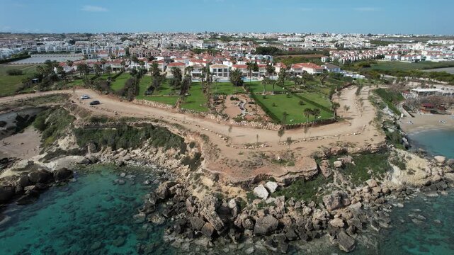 Drone aerial view of holiday houses and green space  by the coast. The setting includes water and rocky areas with a bright sky overhead. Protaras holiday resort Cyprus
