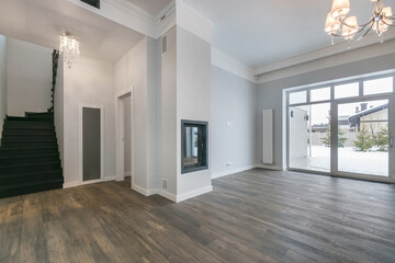 Obraz premium A bright living room with large windows shows a view of snow outside. A staircase leads to the upper level. The fireplace is on the wall as light fixtures hang from the ceiling.