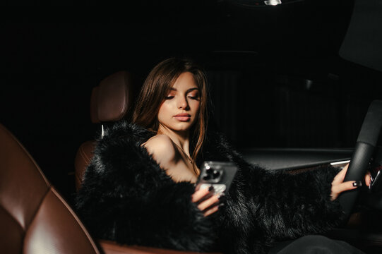 A portrait of a beautiful young, stylish woman, a female model in a fur coat and lingerie, sitting and posing inside an expensive, luxury premium car, holding a phone. Advertising shoot.