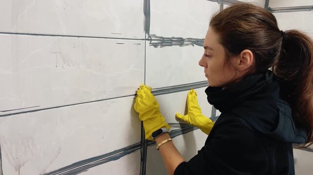 Young European woman in yellow gloves grouting joints between shower tiles. DIY bathroom renovation, waterproofing and hygiene concept, modern home improvement video