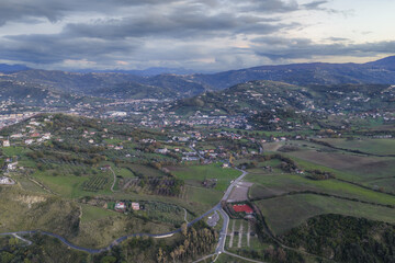 Aerial view of rolling green hills dotted with quaint homes under a vast cloudy sky, a serene landscape stretching into the distant mountains, Agropoli, Campania, Italy.