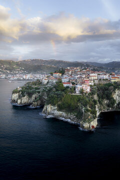 Aerial view of rugged cliffs meet the deep blue sea, topped with sun-kissed buildings and a faint rainbow arching above, Agropoli, Campania, Italy.