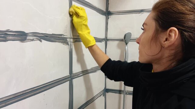 Young European woman in yellow rubber gloves sealing grout lines on shower tiles. DIY bathroom renovation, home improvement, hygiene and waterproofing concept