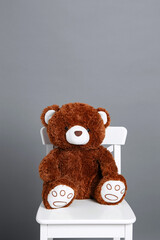 A brown teddy bear sits on a white chair against a gray wall.