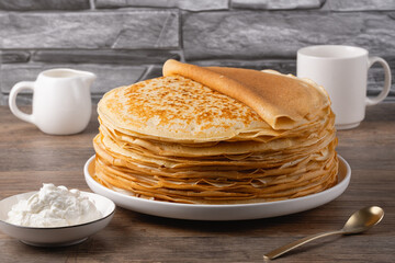 Stack of golden crepes served with sour cream on a wooden table