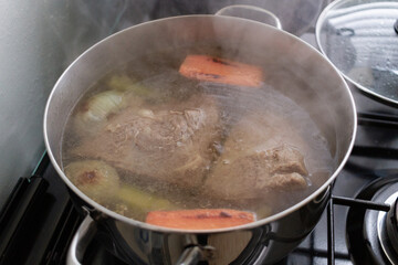 Large stainless steel pot simmering beef, onions, celery and carrot pieces on a gas stove, steam rising as rich homemade broth slowly cooks in a bright family kitchen, bubbles breaking on the surface.