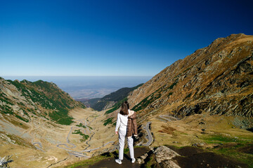 Back view of young woman standing on cliff edge with panoramic mountain landscape of Transfagarasan Highway, Romania. Travel concept