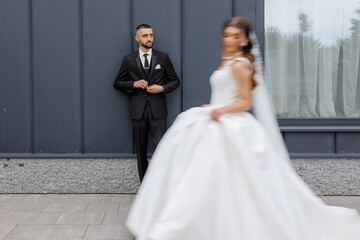 Fototapeta premium Groom adjusts tie while bride walks by in elegant wedding attire at modern venue