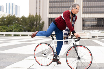 Senior man balancing on fixed-gear bicycle on tiled plaza donning sweater striped scarf glasses