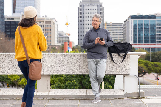 Diverse senior couple standing on rooftop balcony by railing and gym bag while holding smartphones - Powered by Adobe