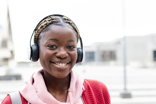 African American woman smiling wearing headphones backpack strap hoodie and jacket in urban plaza
