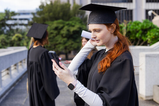 Female graduates wearing gowns checking smartphone and holding diplomas on terrace outside building