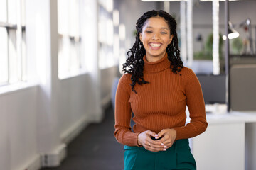 African American woman standing in office corridor wearing rust sweater, teal pants with partitions