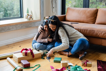 Asian mother and Indian daughter wrapping gifts on living room floor with scissors and ribbons