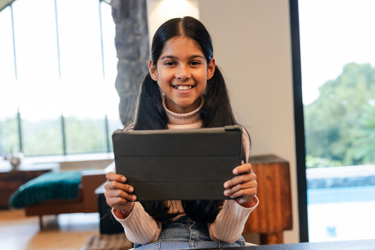 Asian Indian girl sitting on stool in modern living room holding tablet and smiling toward camera