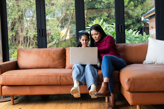 Diverse mother and Indian daughter studying together in home on tan leather sofa with silver laptop