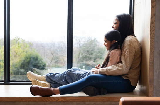 Mother and daughter wearing sweaters embracing on bench gazing through glass window, copy space