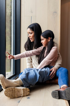 Indian mother cradling child and child pointing through metal frame window on wooden seat at home