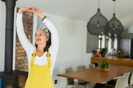 Senior woman stretching arms overhead in kitchen near wood-burning stove, potted plant, copy space