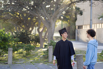 Diverse male friends talking on campus path with posts wearing cap and gown holding rolled diploma