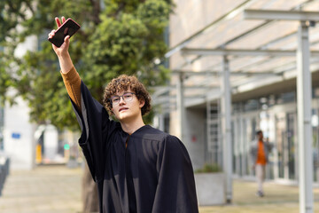 Male graduate student holding smartphone aloft in campus plaza wearing black gown and brown sweater
