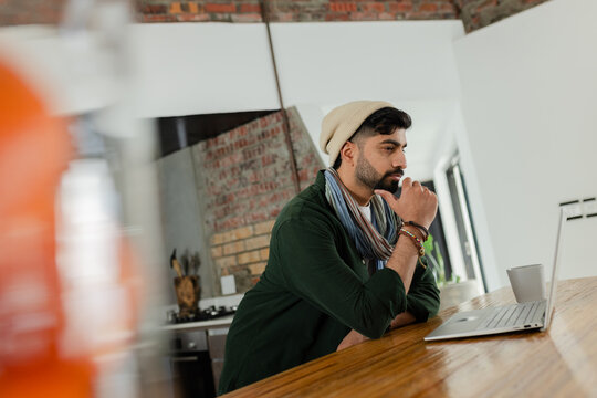 Indian man leaning on wooden kitchen counter, staring at silver laptop with ceramic mug by window