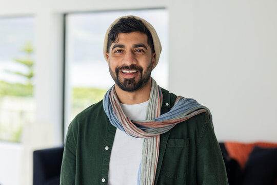 Indian man standing in bright living room wearing beige beanie and striped scarf near window