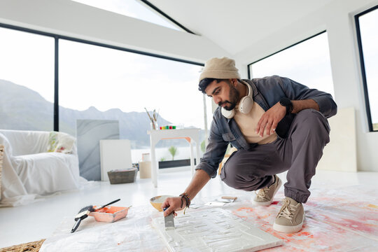 Adult Indian man kneeling on cloth in studio, spreading white medium onto canvas with spatula
