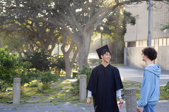 Diverse male friends talking on campus path with posts wearing cap and gown holding rolled diploma