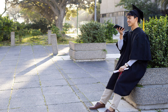 Asian male graduate using smartphone and holding cap plus diploma beside campus planter, copy space