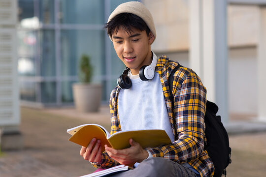 Asian male student sitting on ledge outside campus wearing headphones reading yellow notebook