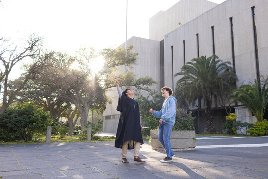 Diverse male friends celebrating graduation in campus courtyard holding diploma and mortarboard cap