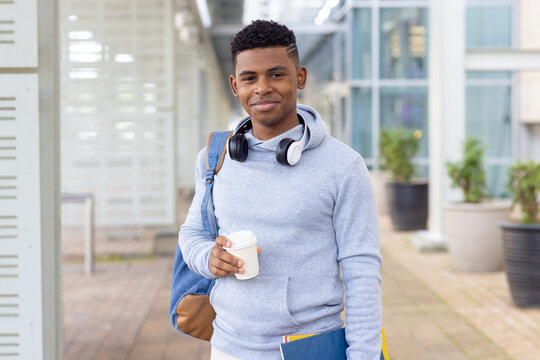 African American man standing on walkway outside glass building, holding coffee cup and notebook
