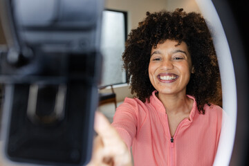 African American woman in pink jacket reaching for smartphone on ringlight stand studio, copy space
