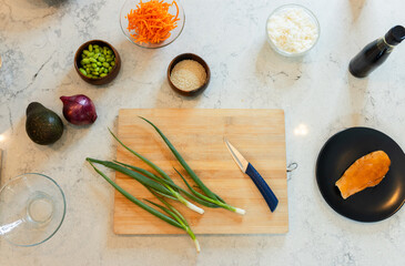 Wooden board holding green onions and blue-handled knife on marble countertop with bowls and salmon