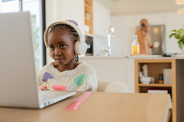 African American girl wearing headphones using laptop at table while parent holding mug, smartphone