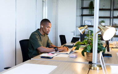 African American man sitting at office desk typing on laptop wearing earbuds and using notebook