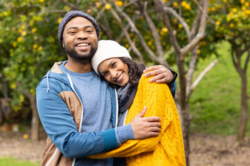 Diverse couple hugging and smiling while standing in fruit tree orchard wearing beanies and scarf