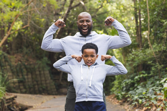 African American father and son flexing muscles while standing on paved forest trail with bench