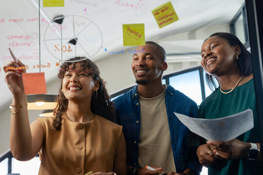 Diverse coworkers collaborating and writing on glass whiteboard at office with sticky notes