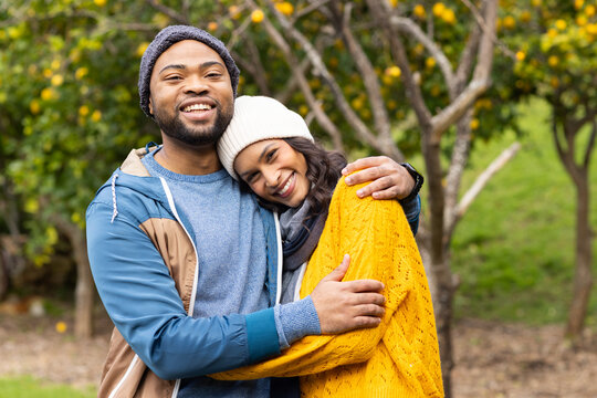 Diverse couple hugging and smiling while standing in fruit tree orchard wearing beanies and scarf