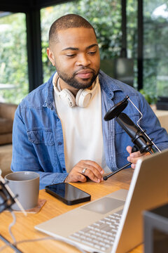 Black adult male podcaster sitting at home office adjusting black mic with pop filter near laptop