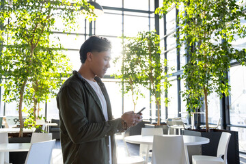 Asian man standing by office lounge tables and planters using smartphone beside windows, copy space