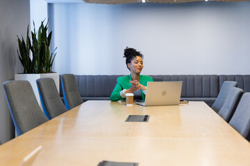 African American woman wearing green blazer sitting at boardroom table, using silver laptop