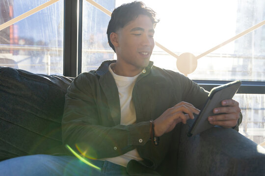 Asian man sitting on dark grey couch in modern lounge area tapping tablet by metal-frame window - Powered by Adobe