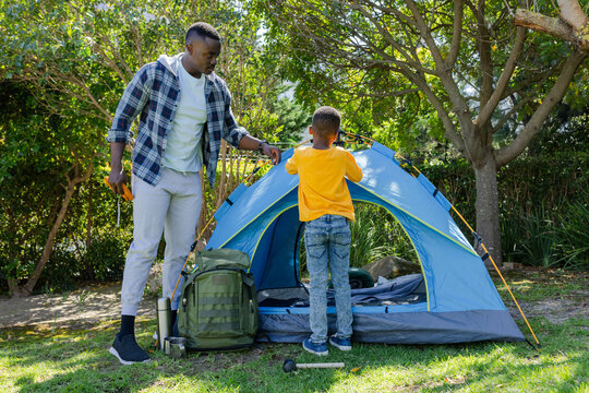 African American man and child boy pitching blue dome tent on grassy patch, holding orange mallet