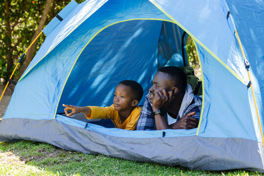 African American father and school-age son lying in blue tent pointing toward woods with backpack