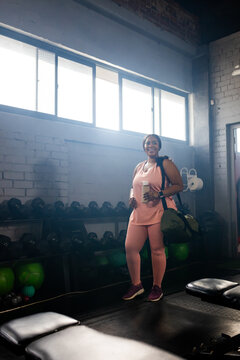 African-American woman wearing pink gear at gym by dumbbell rack with bottle and duffel, copy space