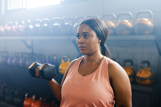 African American woman lifting hex dumbbell near shoulder in coral top at gym with kettlebells