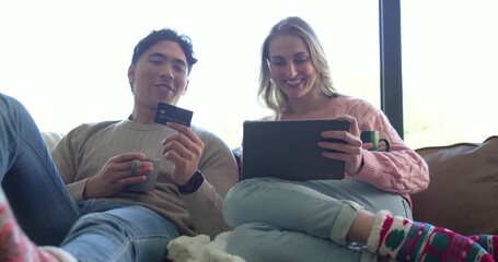 Diverse couple holding mugs and shopping online on tablet on couch after seeing credit card